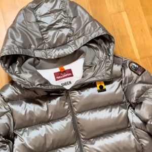 Silver pufferjacka Parajumpers - Säljer en snygg silverfärgad pufferjacka från Parajumpers med huva och coolt märke på ärmen. Jackan har dragkedja framtill och quiltad design som ger en modern vibe. Perfekt för dig som vill sticka ut med stil i vinter.