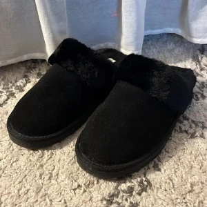 Svarta fluffiga slippers i storlek 40/41 - Mysiga svarta slippers från lager 157 med fluffig fuskpäls runt hela insidan och ovandelen. De är helt oanvända. De har en platt sula i syntet och är perfekta att glida runt i hemma. Enkel slip-on-modell med rund tå och mjuk känsla.