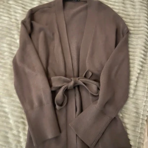 Brun stickad kofta med knytband från LAGER 157 - Säljer en brun väldigt populär , stickad kofta från 157 i storlek S. Koftan har lång ärm och ett matchande bälte i midjan för en chill look. Materialet är mjukt och skönt, perfekt för lager-på-lager. Enkel design utan knappar eller dragkedja. Helt oanvänd