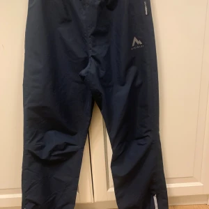 Mörkblå regnbyxor, Stay dry Jr, McKinley, stl 146/152 - Mörkblå regnbyxor från McKinley med elastisk midja och logga på benet. Stl 146/152. Byxorna är tillverkade i ett slitstarkt syntetmaterial och har reflexdetaljer vid benslut. Perfekta blöta dagar utomhus. Mycket sparsamt använda. 