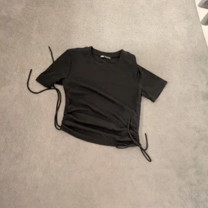 Svart croppad topp med snörning - Svart croppad topp från Bershka med korta ärmar och snygg snörning på båda sidorna. Toppen har en rund halsringning.