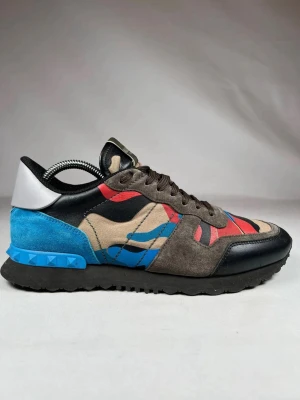Valentino Rockrunner sneakers camo - Säljer ett par Valentino Rockrunner sneakers med färgstarkt camouflagemönster i blått, rött, beige och svart. Skorna har detaljer i mocka och skinn, svart platt sula och snörning. Bakpartiet har blå och vit kontrast med nitar. Perfekt för dig som gillar statement-skor. Skriv om ni har frågor eller funderingar!😊