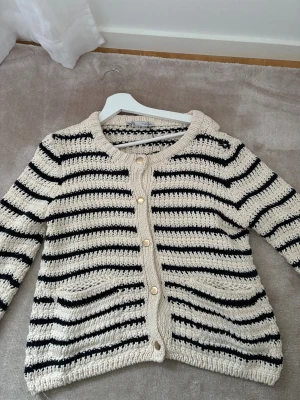 Randig stickad kofta från Zara - Snygg stickad kofta från Zara i storlek L men sitter som en S eller M Koftan är beige med svarta ränder, har rund hals, guldfärgade knappar och två fickor framtill. Materialet är grovstickad bomull som ger en chill och trendig vibe. 