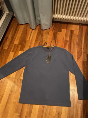 Blå långärmad tröja från Hugo Boss - Snygg blå långärmad tröja från BOSS med diskret broderad logga på bröstet och orange detalj vid halsen. Tröjan har en klassisk rund hals och är gjord i mjuk bomull som känns skön mot huden. Perfekt för dig som gillar stilrena och enkla plagg. Tröjan är helt oanvänd med lappen kvar. Nypris 800kr.