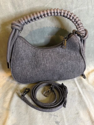 Grå axelväska med flätat handtag NY - Snygg grå axelväska med coolt flätat handtag i rep och matchande justerbar axelrem. Väskan har en mjuk, rundad form och är tillverkad i ett slitstarkt tygmaterial. Dragkedja upptill och metall-detaljer ger en modern vibe. Perfekt storlek för det viktigaste. Mått se bild⬆️