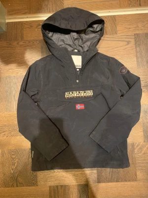 Svart vindjacka från Napapijri - Säljer en snygg svart vindjacka från Napapijri med huva och halv dragkedja. Jackan har en stor ficka framtill med Napapijri-logga och norsk flagga, samt två sidofickor med dragkedja. Den har även en dragkedja på vänstersida från under armhålan och neråt. Dragskedjan framtill saknar själva hållaren men väldigt lätt att fixa en ny. Du kan även spänna åt jackan längst ner. Perfekt för vintern. Vid frågor, hör av dig!