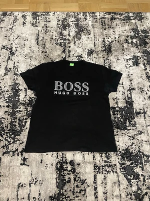 Svart BOSS t-shirt med logga XXL - Svart t-shirt från Hugo Boss i storlek XXL med stor BOSS-logga framtill. Loggan har ett coolt mönster med små BOSS-texter inuti bokstäverna. Klassisk rund hals och korta ärmar. Materialet är mjuk bomull som känns skönt mot huden.