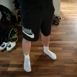 Svarta Nike shorts med logga - Svarta shorts från Nike med stor vit logga på vänster ben. De har elastisk midja med snörning och två sidofickor. Perfekta för träning eller chill, tillverkade i mjuk bomullsmix för skön känsla. Klassisk loose fit som ger en avslappnad look.