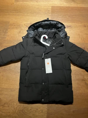 Svart dunjacka från Canada Goose - Svart dunjacka från Canada Goose med huva och avtagbar pälsdetalj. Jackan har klassisk patch på ärmen, dragkedja och knappar framtill. Fylld med dun och har en loose passform, perfekt för kalla vinterdagar. Snygg och tidlös design som håller dig varm. Storlek M och kommer med prislapparna.