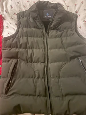 Svart pufferväst från DI-BYE - Svart pufferväst från DI-BYE med hög krage och två fickor med dragkedja. Västen har quiltad design och är ärmlös, perfekt att slänga över en hoodie eller tröja. Stängs med dragkedja framtill och har en clean look.