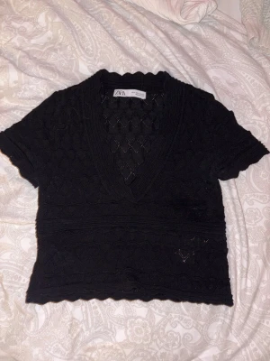 Svart stickad topp från Zara - Supersnygg svart stickad topp från Zara med v-ringad hals och korta ärmar. Toppen har ett genombrutet mönster och vågiga kanter som ger en extra cool vibe. Perfekt att styla med jeans eller kjol för en trendig look.