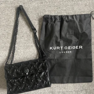 Svart quiltad axelväska från Kurt Geiger - Svart axelväska från Kurt Geiger med glansigt quiltat skinn och coola detaljer i form av små fåglar och en unik handformad låsdetalj. Väskan har en kedjerem i metall och kommer med dustbag. Perfekt storlek! 