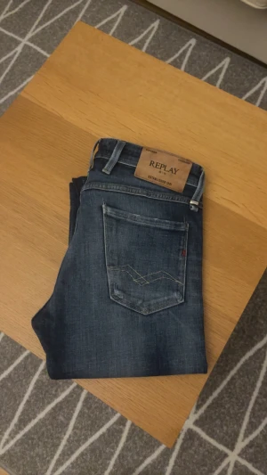 Replay Anbass slim fit jeans blå - Snygga Replay Anbass jeans i slim fit med klassisk femficksdesign. Färgen är mörkblå med slitningar och tvättade detaljer för en cool look. Byxorna har dragkedja och knapp, samt detaljerad söm på bakfickan. Perfekt för dig som gillar stilrena jeans med lite edge.