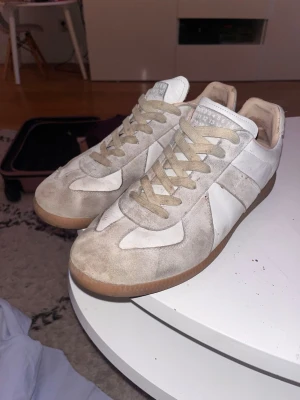 Maison Margiela sneakers vit/grå - Maison Margiela  sneakers i vit och ljusgrå färg med beige snörning och gummisula. Klassisk rund tå och platt sula, tillverkade i skinn och mocka. Snygg retrostil med ikonisk etikett på plösen. Perfekta för dig som gillar minimalistisk design.