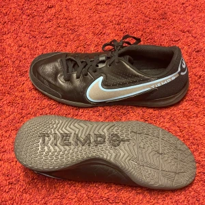 Nike Tiempo Legend 9 Academy IC Renew - Svart/Grå  - Nike Tiempo Legend 9 Academy IC Renew inomhusskor i svart färg med ljusblå och guldgrå detaljer. Dessa skor är designade för att ge bra bollkontroll och ökad precision. Yttersulan i gummi är utformad för grepp i flera riktningar på inomhusytor. Ovandelen har upphöjda texturer med mjuka skumkuddar för exakt dribbling, passning och skott.   Ovandelen är i KALVSKINN vilket formar sig efter foten och har god andningsförmåga. Mycket bra skick, i nära nyskick. 