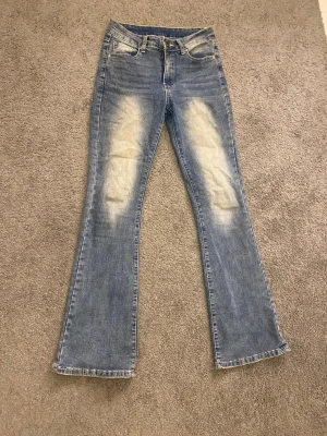 Blå bootcut jeans med detaljerade fickor - Snygga blå bootcut jeans med ljusa slitningar framtill och bak. Baksidan har två fickor med dekorativa sömmar och silverfärgade knappar. Jeansen har klassisk femficksdesign och är tillverkade i jeansmaterial med normal passform. Storlek S men passar en Xs oxå