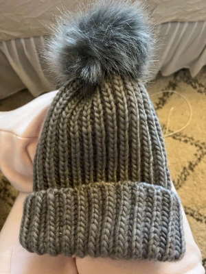 Grå stickad mössa med pälsboll - Säljer en chunky grå stickad mössa med en stor fluffig pälsboll på toppen. Mössan är ribbstickad och har en bred uppvikt kant. Materialet är 100% polyester och den är perfekt för kalla dagar.