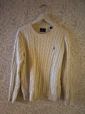 GANT tröja  - Säljer en vit kabelstickad tröja från GANT. Tröjan är gjord av premium bomull och har rund hals, ribbade muddar och GANT-logga broderad på bröstet. Axelbredd 42cm, längd 62cm. JAG SKICKAR INGA BILDER NÄR JAG BÄR PLAGGET:) obs se defekt på sista bilden. !!