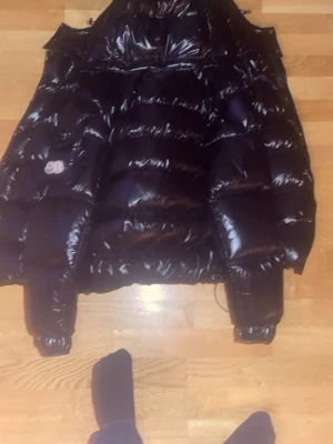 Svart Moncler pufferjacka - Säljer en svart pufferjacka från Moncler med glansig finish och klassisk logga på fickan. Jackan har quiltad design, dragkedja och långa ärmar. Insidan har en cool serietidningsetikett. Perfekt för dig som vill ha en varm och snygg vinterjacka.
