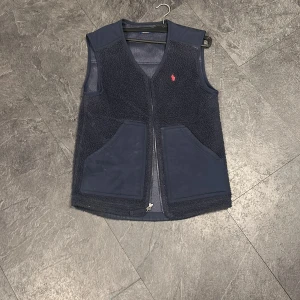  teddyväst från Polo Ralph Lauren - Snygg mörkblå teddyväst från Polo Ralph Lauren med klassisk röd logga på bröstet. Västen har två stora fickor fram och dragkedja hela vägen. Materialet är mjuk fleece och polyester, perfekt för lager-på-lager. Stilren och enkel design utan ärmar.