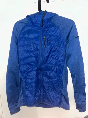 Peak Performance jacka - Blå pufferjacka från Peak Performance med huva och hel dragkedja framtill. Jackan har quiltade paneler, sidofickor med dragkedja och broderad logga på ärmen. Materialet är syntet och polyester, perfekt för kyliga dagar. Snygg och sportig look. Pris kan diskuteras vid snabbaffär.