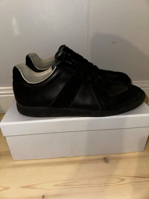Maison Margiela svarta sneakers - Maison Margiela sneakers i helsvart med rund tå och platt sula. Ovandelen är en mix av skinn och mocka, med svarta snören och vit insida. Klassisk siluett med diskret branding på tungan och undersidan av sulan. Perfekt för dig som gillar minimalistisk och stilren design.