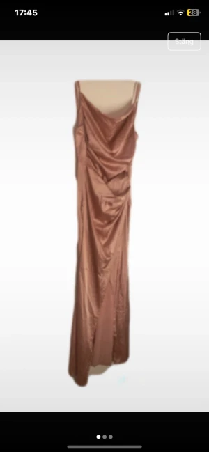 Champagnefärgad långklänning i satin - Snygg långklänning (balklänning) i champagnefärgad satin med tunna axelband och draperad front. Klänningen har en elegant, släpande siluett och ett mjukt, glansigt material som ger en lyxig känsla. Perfekt för dig som vill sticka ut med en stilren och trendig look.