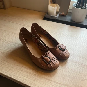 Bruna pumps med spänne från Gabor - Snygga bruna pumps från Gabor med låg klack och rund tå. Skorna är gjorda i skinn och har ett dekorativt spänne framtill samt veckade detaljer. Så söta!! Storlek UK 5 eller 38! 