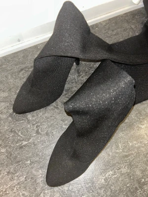 Svarta glittriga stövletter med klack - Svarta stövletter med glittrigt tyg och spetsig tå. Skorna har en smal klack och är tillverkade i stretchigt syntetmaterial som sitter tight runt benet. Perfekt för dig som vill ha en edgy och trendig look med extra glitter. klackarna är köpta från italien. ✨