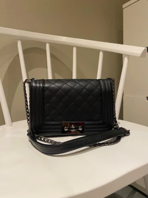 Svart quiltad axelväska med kedja - Snygg svart axelväska i quiltat skinn med silverfärgad kedjerem och stilren låsdetalj framtill. Väskan har en klassisk rektangulär form och passar perfekt för dig som gillar en tidlös men trendig look. Bandet går att ha i olika längder. 