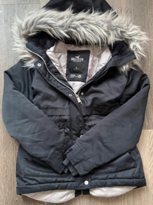 Svart vinterjacka från Hollister - Snygg svart vinterjacka från Hollister med huva och grå fuskpäls runt kanten. 