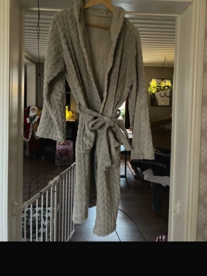 Grå morgonrock med snörning - Mysig grå morgonrock i fleece med snyggt mönstrad yta och huva. Västen har ett knytband i midjan för att justera passformen och ger en chill vibe. Perfekt för att hålla sig varm och bekväm under kyliga dagar.