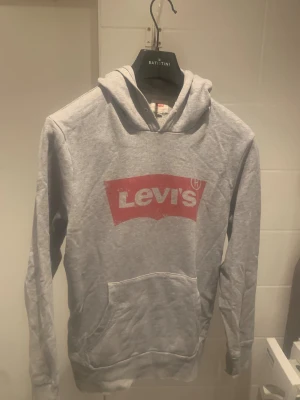 Grå Levi's hoodie med röd logga - Säljer en grå hoodie från Levi's med stor röd logga på bröstet. Tröjan har huva, lång ärm och en klassisk magficka framtill. Perfekt för dig som gillar streetwear och vill ha en enkel men snygg look.