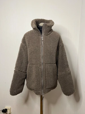 Brun teddyjacka från H&M XS - Mysig brun teddyjacka från H&M i storlek XS. Jackan har hög krage, två stora fickor framtill och stängs med dragkedja. Perfekt för kyliga dagar när du vill vara både varm och snygg. Oversized passform för en avslappnad vibe. Har redan en i mörkbrun och svart. 