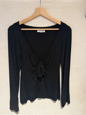 Svart långärmad topp med knyt - Svart långärmad topp från Design by Si i storlek M. Modellen har djup v-ringning och snygg knytning framtill som ger en cool detalj. Tunn och mjuk bomullskänsla, perfekt till jeans eller kjol. Enkel och stilren look med figurnära passform.