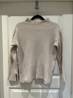 Beige ribbad stickad tröja Filippa K - Snygg beige stickad tröja från Filippa K med ribbad struktur och coola diagonala detaljer. Tröjan har långärmade ärmar med slits vid mudden och en högre hals som ger en chill vibe. Perfekt för dig som gillar stilrena och mjuka plagg.