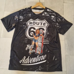 Svart Route 66 t-shirt med tryck - Cool svart t-shirt med stort Route 66-tryck och retro bensinstationsmotiv på både fram- och baksidan. T-shirten har korta ärmar och en rund hals. Materialet känns lätt och syntetiskt, perfekt för dig som gillar äventyr och statement-plagg.