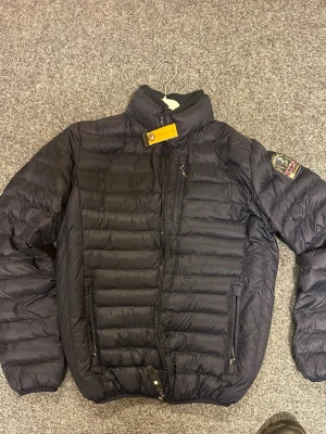 Svart dunjacka från Parajumpers - Säljer en svart dunjacka från Parajumpers med blått foder, gul detalj vid kragen och patch på ärmen. Jackan har dragkedja, hög krage och två fickor med dragkedja. Perfekt för kalla dagar och riktigt snygg streetstil.