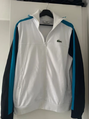 Lacoste zip up tröja - Snygg långärmad tröja med dragkedja från Lacoste i vitt. Jackan har sidofickor och broderad Lacoste-logga på bröstet. På ryggen finns en diskret broderad signatur och tennisspelare. Riktigt stilren och snygg tröja! Tröjan är i Stl S men stor i storlek. Jag är vanligtvis mellan M och L och passar mig!