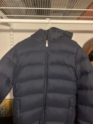 Mörkblå pufferjacka med huva från Sail Racing - Säljer en mörkblå pufferjacka med huva och dragkedja framtill. Jackan har breda, quiltade sektioner och en clean look utan synliga loggor. Perfekt för kalla dagar och har en snygg, enkel design som funkar till de flesta stilar.