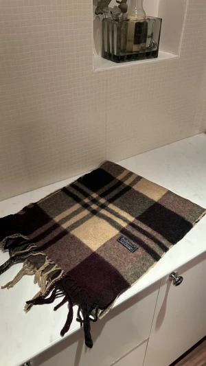 Rutig ullväst från Burberrys - Säljer en klassisk rutig väst från Burberrys i mjuk ull. Färgerna är beige, brun, mörkgrå och vinröd med typiskt Burberry-mönster. Västen har fransar längs nederkanten och en snygg etikett framtill. Perfekt för dig som gillar tidlös stil och vill ha något extra.