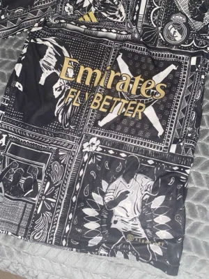 Real Madrid Bellingham fotbollströja - Säljer en unik Real Madrid fotbollströja med Bellingham och nummer 5 på ryggen. Tröjan har svart och guld färg med grafiska mönster och detaljer som klubbmärke och Emirates-tryck. Materialet är lätt polyester med Aeroready-teknologi för extra komfort.
