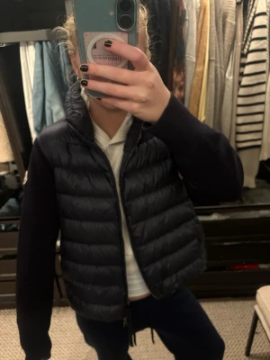 Mörkblå dunväst från Moncler - Snygg mörkblå dunväst från Moncler med stickade ärmar och quiltad framsida. Västen har hög krage, dragkedja och Moncler-logga på ärmen. Perfekt för lager-på-lager och har en clean, sportig look. Materialet är dun och stickad polyester. Köptes för runt 7500 kr men säljs nu för endast 3000. Skriv om ni har fler frågor❤️💥