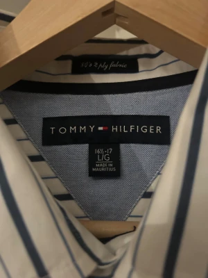 Randig skjorta från Tommy Hilfiger - Snygg vit skjorta med blåa vertikala ränder från Tommy Hilfiger. Klassisk krage, långärmad och med knappar framtill. Materialet är mjuk bomull och på bröstfickan finns den ikoniska Hilfiger-loggan broderad. Perfekt för dig som gillar stilrena och fräscha plagg.