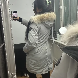 Canada goose jacka - Säljer en snygg ljusgrå parkas från Canada Goose med fluffig päls på huvan. Jackan har stora svarta knappar, dragkedja och två rejäla fickor framtill. Insidan är svart och jackan är perfekt för kalla vinterdagar. Huvan är fodrad med mjuk päls och jackan har lång ärm. Äkta päls, den gör mig jättevarm på vintern!!! Kan mötas upp i Stockholm, går ner i pris för snabbaffär❤️