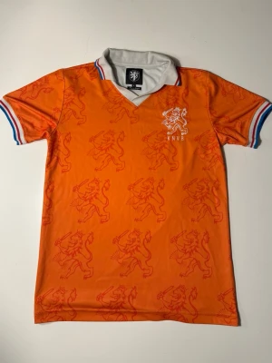Nederländska Fotbollströja 1994 - Tryck på tröjan kan önskas! Tröjan har den klassiska orange färgen, ett snyggt mönster med lejon över hela tyget och KNVB-emblemet på bröstet. Kvaliteten är riktigt bra mjukt, hållbart material och fina detaljer i krage och ärmslut. Perfekt för samlare, fotbollsälskare eller dig som vill sticka ut med en retroklassiker.