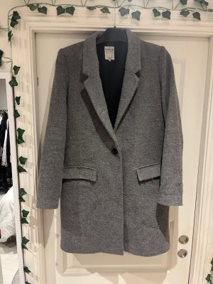 Grå kappa från Zara  - Snygg grå kappa från Zara Trafaluc med klassisk krage och enkel knäppning framtill. Kappan har två fickor med lock och är helfodrad. Perfekt för dig som gillar stilrena och tidlösa plagg med en modern touch.
