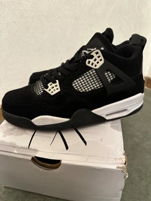 Nike Air Jordan 4 Retro svart/vit - Säljer ett par Nike Air Jordan 4 Retro i svart och vitt. Skorna har meshpaneler, svarta snören och ikoniska plastdetaljer vid snörningen. Ovandelen är i mocka och textil, med en vit och svart sula som ger en clean kontrast. Perfekta för dig som gillar streetwear och sneakers med attityd.