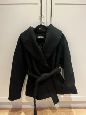Svart kappa med bälte från Gina Tricot - Snygg svart kappa från Gina Tricot i storlek XS. Jackan har bred krage, knytbälte i midjan och långa ärmar. Materialet är mjukt och känns lyxigt, perfekt för kyliga dagar. Enkel och stilren design som funkar till många outfits. Använd max 5 gånger, storlek xs passar mig som har s!