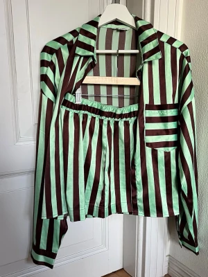 Randigt pyjamas set - Skjorta och kort byxa med resår i satin från Monki. Breda vertikala ränder i mintgrönt och mörkbrunt. Kan användas som pyjamas eller på dagen varje plagg för sig eller i set för en coolt dressad look. Fräsch, obetydligt använd. 